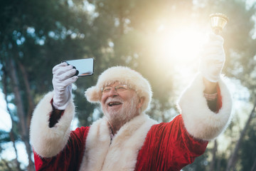 From below joyful man in costume of Santa Claus ringing bell and taking selfie with mobile phone on blurred nature background