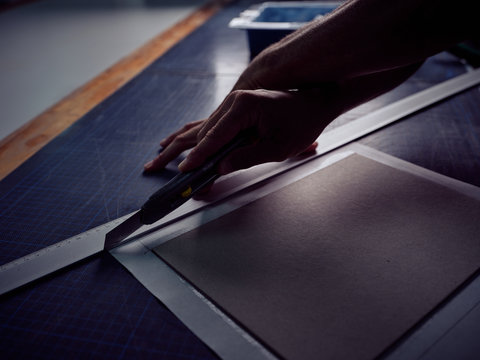 From above crop bookbinder cutting paper with knife and ruler on blue checkered mat in workshop