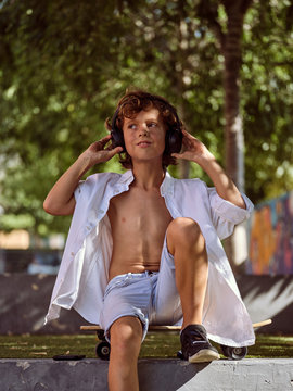 Casual pensive boy in headphones sitting on skateboard while relaxing in skatepark on sunny summer day and looking away