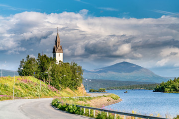 Old church at the left side of Umea river, heavy rainy clouds and mountain range at background. E12 european road curves at the foreground. Summer day with clouds and sun. Tarnaby village, Lapland