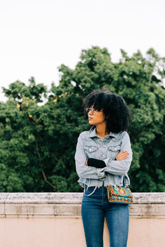 Young trendy African American woman in jeans and crop top sitting on stone parapet