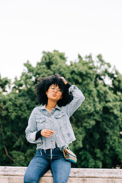 Young trendy African American woman in jeans and crop top sitting on stone parapet