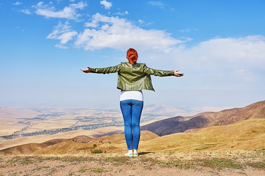 Beauty woman outdoors enjoying nature. Image of a girl in a field standing in the distance with her back to the camera and her arms outspread in celebration of a beautiful sunny summer day and freedom