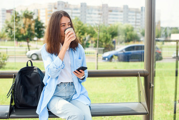 Charming hipster girl waiting for bus or tram on public transport station in the morning with cup of coffee and uses smart phone and internet to monitoring transport through the app.