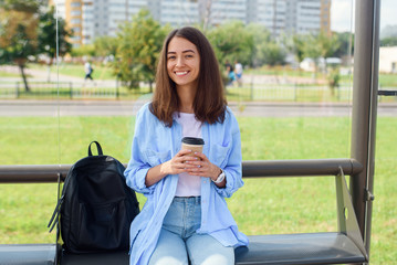 Charming hipster girl waiting for bus or tram on public transport station in the morning with cup of coffee and uses smart phone and internet to monitoring transport through the app.