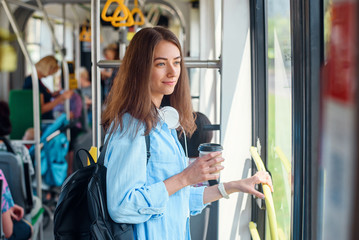 Young woman in blue shirt rides in the modern tram or bus with cup of coffee to go.