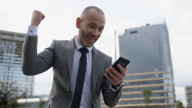 Happy bearded businessman in suit cheering celebrating success and achievement looking at his smart phone. Handsome professional man receiving good news in business on phone, happy emotions outdoors