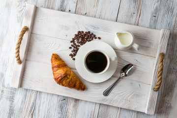 A white Cup of coffee with a croissant on a light wooden tray top view