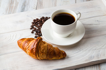 A white Cup of coffee with a croissant on a light wooden tray