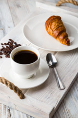 A white Cup of coffee with a croissant on a light wooden tray
