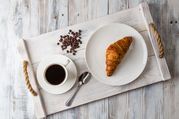 A white Cup of coffee with a croissant on a light wooden tray