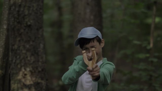 boy in a forest jumps out a tree and takes aim at the camera with a slingshot