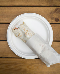 Fototapeta na wymiar bitten doner kebab on white dish on wooden table, flat lay