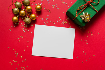 Christmas composition. White blank paper, wrapped gift, toys on red background with golden confetti. new year concept. Greeting card, xmas celebration 2020. Flat lay, top view, copy space, mockup