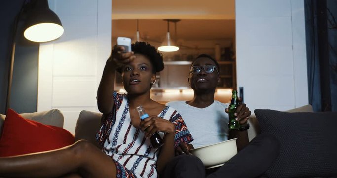 Happy positive young African man and woman smile and talk watching TV, using remote at home with snacks slow motion.