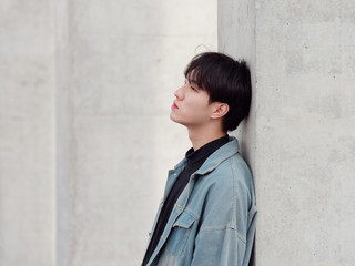 Obraz na płótnie Canvas Portrait of a handsome Chinese young man in jean leaning against wall and looking away with blank expression, has nothing to do just waiting.