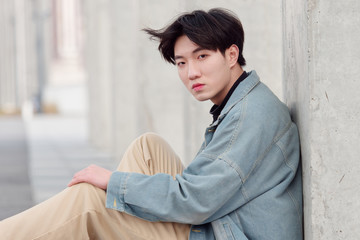 Portrait of a handsome Chinese young man in jeans sitting on ground and looking at camera.