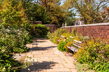 A stone walkway in a garden of flowers with two wooden benches and a brick wall in summer, Pittsburgh, Pennsylvania, USA
