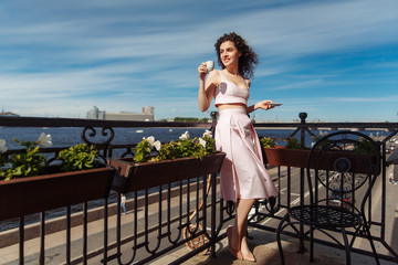 Fototapeta na wymiar Young beautiful girl with curly hair in a pink dress is standing on the balcony and drinking coffee. Happy woman smiling and looking at the Neva River in St. ..Petersburg