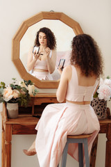 Young happy girl with curly hair does makeup in front of a vintage mirror. A beautiful woman in a pink dress paints her eyes.