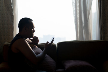 Asian man in armless clothes sitting in room near clear glass window, eyes looking at smart phone