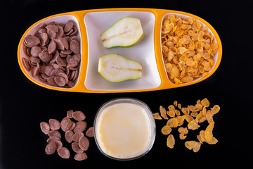Corn choco flakes cereal in yellow tray with milk and pear slices isolated on black gray background, a delicious and healthy  dietary breakfast. Top View.