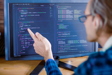 Man freelancer in glasses is writing software, working at home, office. Programmer developer is showing program code script on big computer screen. Geek desktop, workplace concept. Remote work.