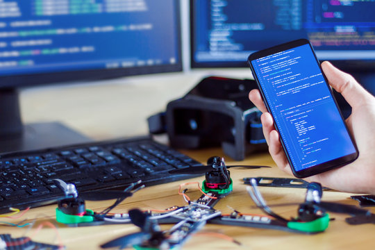 Closeup programmer hand is holding smartphone with program code software on screen for controlling FPV drone. Man developer is building constructing quadcopter from kit with microcontrollers.
