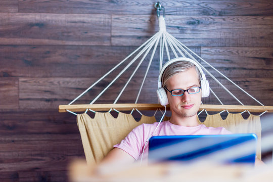 Young man freelancer in headphones is working on laptop while lying in cozy hammock at flat. Programmer developer in glasses is writing program code software. Geek home workplace concept. - Powered by Adobe