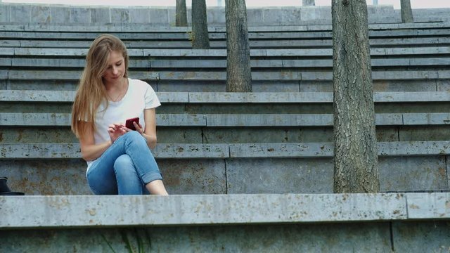 Young beautiful girl in a park holds a smartphone in her hands, writes a message. Leafing through photos on the phone, chatting in social networks with friends. Sits on concrete.