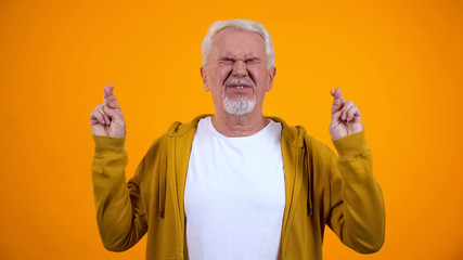 Male in his 50s crossing fingers for luck, hoping for victory, orange background