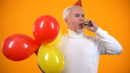 Glad male pensioner using party blower and holding colorful balloons, birthday