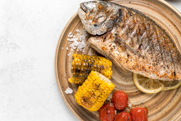 From above grilled fish with lemon served with different vegetables on dishes over white background