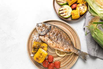From above grilled fish with lemon served with different vegetables on dishes over white background