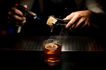 Fototapeta na wymiar Professional bartender serving a brown cocktail melting caramel with a burner above the lemon slice in the dark