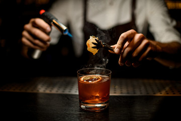 Fototapeta na wymiar Bartender serving a brown cocktail melting caramel with a burner above the lemon slice in the dark