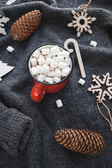 Hot chocolate in a red mug with marshmallow surrounded Christmas decoration on a woolen sweater.