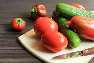 Fresh vegetables with a knife lie on a wooden board. Near the table are two peppers.