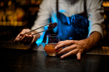 Professional barman adding chilled melting caramel with twezzers to the cocktail glass with ice cubes under blue lights
