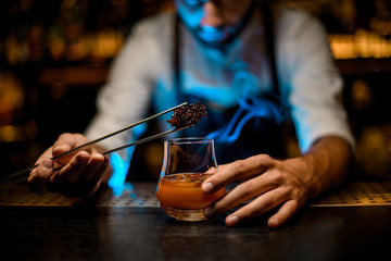 Professional male barman adding chilled melting caramel with twezzers to the cocktail glass with ice cubes under blue lights