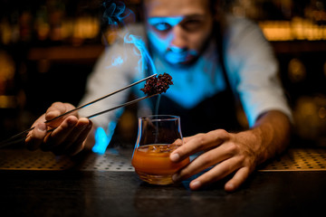 Attractive male bartender adding chilled melting caramel with twezzers to the cocktail glass with ice cubes under blue lights