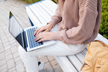 Details hands, legs in jeans and stationery of stylish woman working typing on laptop, freelancer sitting in park on bench working, urban city street style, business center, online education