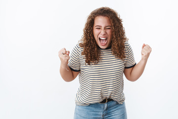 Energized confident curly-haired female showing girl power yelling out loud gather will power fists closed eyes express courage fighting stereotypes break free body-positive movement, white wall