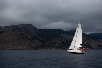 Fototapeta premium Side view of white sail boat on the grey calm water of sea or ocean during cloudy, windy and grey day with yacht team on board and rocks with green landscape on the background