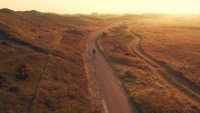 Aerial shot of a young woman cyclist riding a bike on a bike path among sand dunes at golden sunrise in the Texel island in The Netherlands