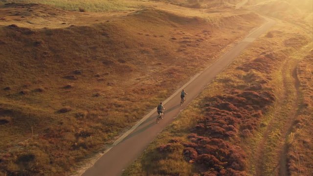 Tracking aerial shot of a young couple riding bikes on a scenic bike path among sand dunes at golden sunrise in the unique landscape of the Texel island in The Netherlands