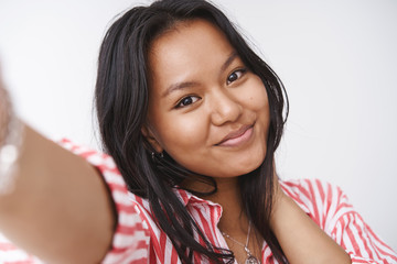 Headshot of charming cute and tender young polynesian woman pulling hand towards camera as taking selfie and looking at camera with lovely gentle smile posing against white background
