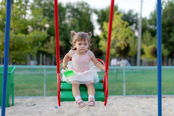 Funny little child, adorable preschooler girl in pretty dress having fun on a swing in the park on summer day