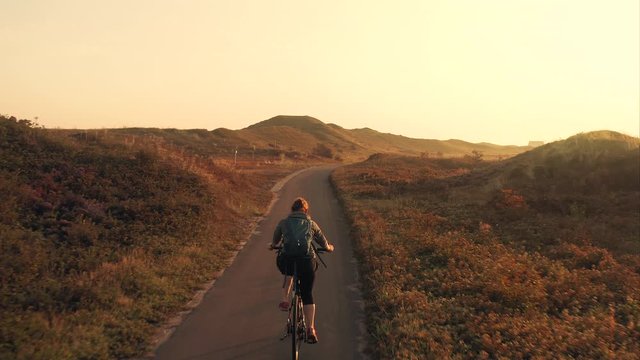 Tracking aerial shot of a young woman cyclist riding a bike on a bike path among sand dunes at golden sunrise in the unique landscape of the Texel island in The Netherlands