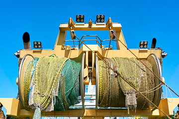Rolled up industrial fishing nets on a winch aboard a yellow fishing boat or trawler against a blue...
